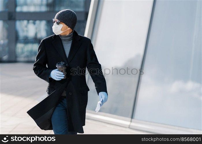 Shot of young man poses outdoor at street, looks aside, wears sunglasses, rubber gloves, medical mask during coronavirus outbreak, drinks hot beverage, tries to avoid public places during epidemic