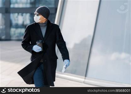 Shot of young man poses outdoor at street, looks aside, wears sunglasses, rubber gloves, medical mask during coronavirus outbreak, drinks hot beverage, tries to avoid public places during epidemic