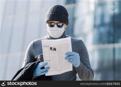 Shot of young man reads article about coronavirus symptoms, wears protective medical mask and rubber gloves, poses at street with coffee, follows rules or quarantine. Covid-19 protection concept