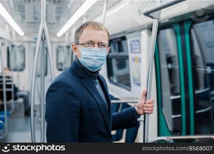 Shot of young man wears spectacles and protective face mask to prevent spreading coronavirus disease or flu epidemic in public transport, poses at empty subway carriage. Public health solution