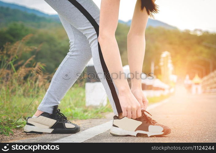 Shot of young woman runner tightening running shoe laces, getting ready for jogging exercise outdoors. Female jogger lacing her sneakers standing on road path before morning run.