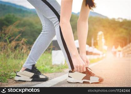 Shot of young woman runner tightening running shoe laces, getting ready for jogging exercise outdoors. Female jogger lacing her sneakers standing on road path before morning run.