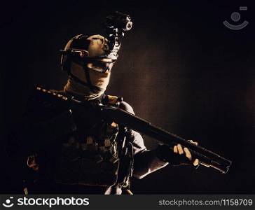Shoulder portrait of army elite troops soldier, anti-terrorist tactical team wit shotgun, helmet with thermal imager, hiding face behind mask, armed rifle with optical scope, studio shoot on black. Army elite troops soldier low key studio portrait