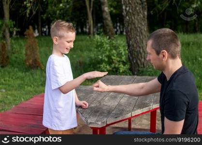 show hands, play with dad, family weekend, white t-shirt, blonde hair, sit on a bench, play in the park. A cute boy is playing a game with his dad. shows signs with his hands
