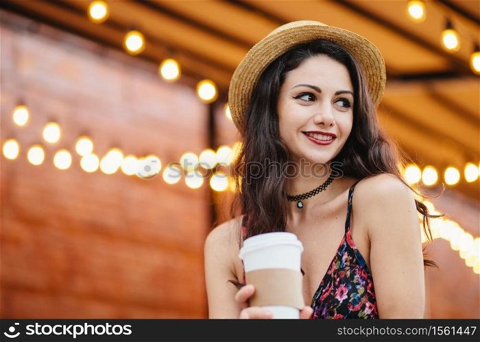 Shy brunette female with dark eyes and red lips wearing straw hat and dress, holding takeaway coffee or tea looking with smile aside being happy to notice her boyfriend, resting at outdoor cafe