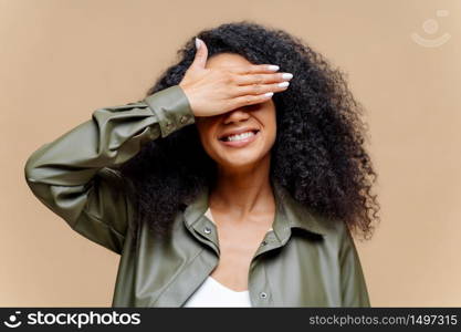 Shy cheerful young Afro woman covers eyes with palm, has toothy smile, hides face, has curly hairstyle, dressed in stylish clothes, isolated over brown background, waits for surprise or gift
