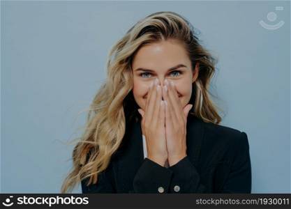 Shy cute blonde girl wearing black coat covering mouth with both of her hands, filled with happiness and joy, can not contain her emotions while standing next to blue background. Surprise concept. Shy blonde girl covering her mouth with hands and feeling excited