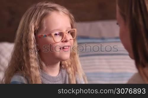 Shy cute little girl in glasses with blonde long hair and blue eyes telling story to mom and kissing her with love. Happy loving family sharing their secret and supporting each other.