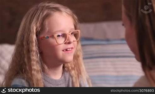 Shy cute little girl in glasses with blonde long hair and blue eyes telling story to mom and kissing her with love. Happy loving family sharing their secret and supporting each other.