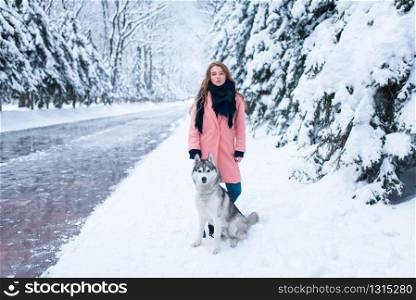 Siberian husky sitting near young woman, snowy forest on background. Cute girl with charming dog. Siberian husky sitting near young woman