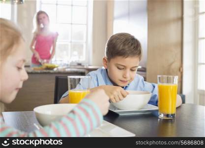 Siblings having breakfast at table with mother preparing food in background