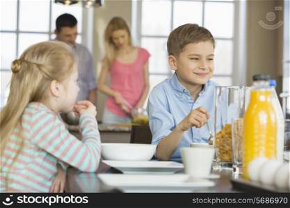 Siblings having breakfast at table with parents cooking in background