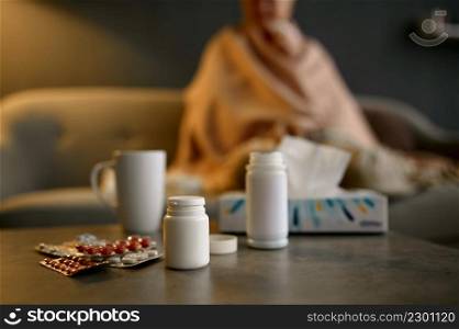 Sick aged woman feeling unwell wrapped in warm quilt. Focus on pills for medical treatment on table. Sick elderly woman and pills for treatment