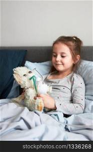 Sick child recovering in bed. Little girl playing by applying medical inhalation treatment with nebuliser to her teddy bear