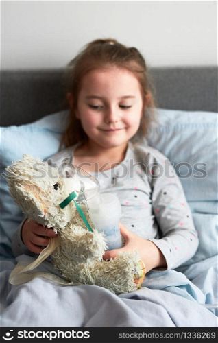 Sick child recovering in bed. Little girl playing by applying medical inhalation treatment with nebuliser to her teddy bear