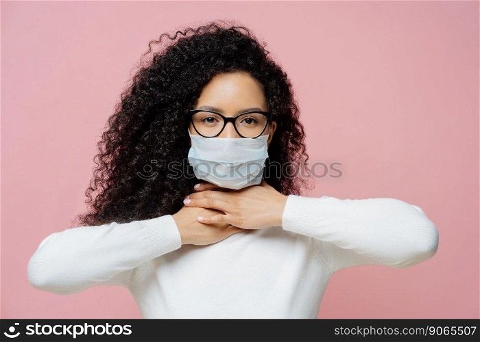 Sick curly haired woman touches neck, suffers from suffocation and shortage of breathing, wears medical mask to avoid virus infection, isolated on pink background. Coronavirus, health care concept