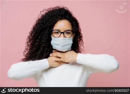 Sick curly haired woman touches neck, suffers from suffocation and shortage of breathing, wears medical mask to avoid virus infection, isolated on pink background. Coronavirus, health care concept