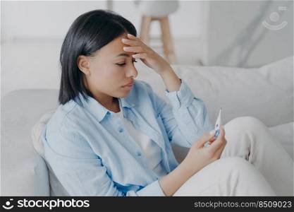 Sick hispanic young woman is measuring temperature at home. Girl is sitting on sofa and looking at the display of electronic thermometer. Headache from coronavirus or influenza. Sick leave concept.. Sick young woman is measuring temperature at home, looking at the display of electronic thermometer.