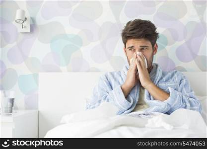 Sick man blowing his nose while sitting on bed at home