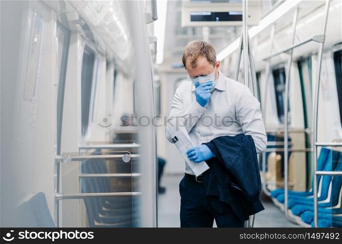 Sick man commuter sneezes constantly, has symptoms of coronavirus, wears surgery medical mask and gloves, holds jacket and newspaper, stands in public transport, has responsible social behavior