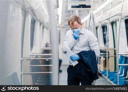 Sick man commuter sneezes constantly, has symptoms of coronavirus, wears surgery medical mask and gloves, holds jacket and newspaper, stands in public transport, has responsible social behavior