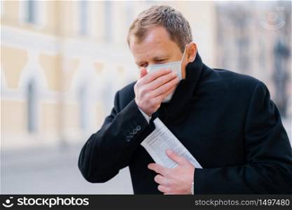 Sick man coughs, covers mouth with palm, wears medical mask, has symptoms of allergy, flu, influenza or coronavirus, walks outdoor, holds newspaper, feels unwell. Covid-19, quarantine concept