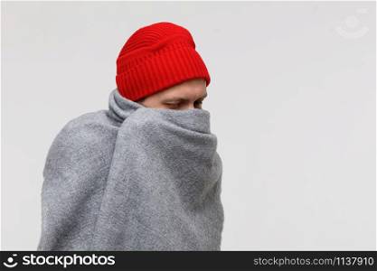 Sick man in red beanie wrapped in warm grey plaid, suffering from cold, virus, fever, looking down, isolated on grey background. Flu season.