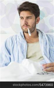 Sick man with thermometer in mouth sitting on bed