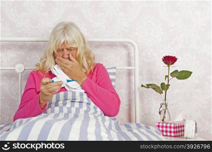 Sick woman sitting in bed looking worried at thermometer with tissues, a rose and washcloth
