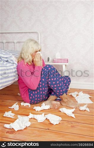Sick woman with pink pajama and tissues sitting on floor against bed