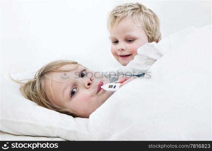 Sick young boy measuring his fever with a thermometer in bed, whilst his younger brother is trying to cheer him up
