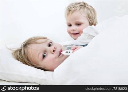 Sick young boy measuring his fever with a thermometer in bed, whilst his younger brother is trying to cheer him up