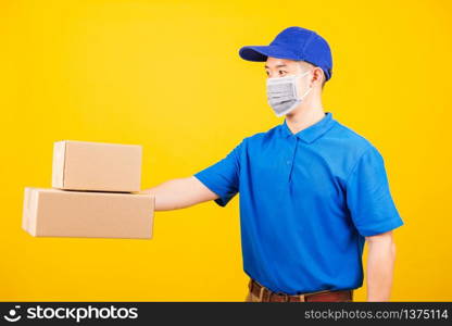 Side of Asian young delivery worker man in blue t-shirt and cap uniform wearing face mask protective giving cardboard boxes under coronavirus or COVID-19, studio shot isolated yellow background