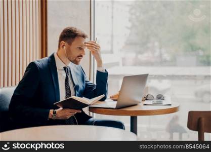 Side photo of successful male company worker in earphones working online from coffee shop, watching business video on laptop computer while sitting next to large bright window in formal stylish suit. Side photo of successful business person busy working from coffee shop