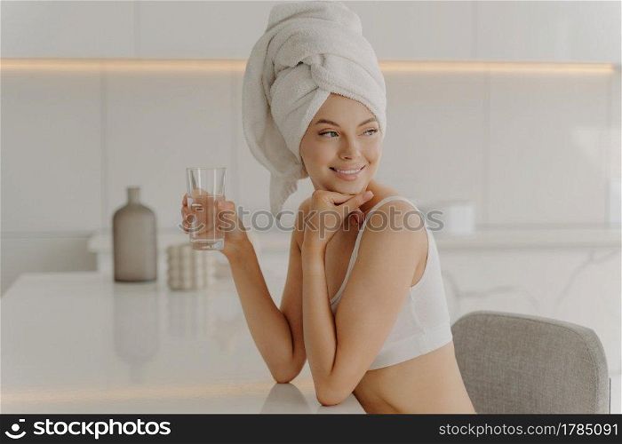 Side portrait of beautiful caucasian female sitting in kitchen after morning refreshing shower with head wrapped in bath towel, drinking mineral water and smiling. Healthy lifestyle concept. Side portrait of beautiful caucasian female sitting in kitchen with glass of water