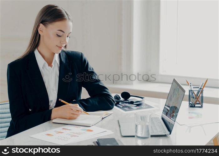 Side portrait of smiling concentrated young caucasian bussiness woman in black formal suit writing in note book while sitting by big white desk with window in light minimalistic background.. Bussiness woman sitting by desk and writing in note book in front of laptop