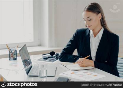 Side portrait of smiling concentrated young caucasian bussiness woman in black formal suit writing in note book while sitting by big white desk with window in light minimalistic background.. Bussiness woman sitting by desk and writing in note book in front of laptop