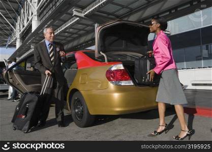Side profile of a businesswoman loading her suitcase on a car trunk with a businessman standing in front of her