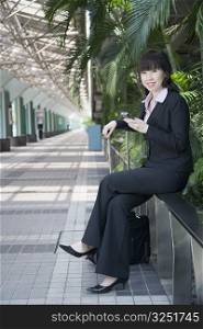 Side profile of a businesswoman sitting on a railing and holding a mobile phone
