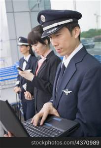 Side profile of a female cabin crew holding a mobile phone and standing with two pilots at an airport