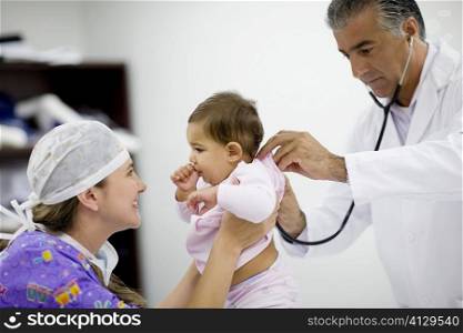 Side profile of a female doctor playing with a baby girl and a male doctor examining her