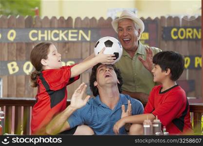 Side profile of a girl balancing a soccer all on her father&acute;s forehead with her brother and grandfather in a restaurant