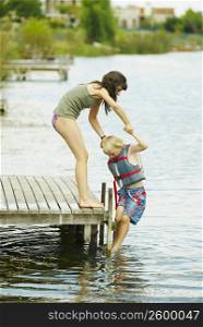 Side profile of a girl standing on a pier and helping her brother out of a lake
