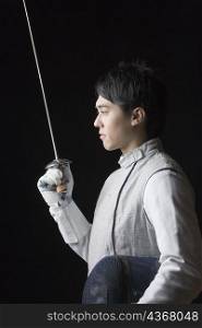 Side profile of a male fencer holding a fencing foil and a fencing mask