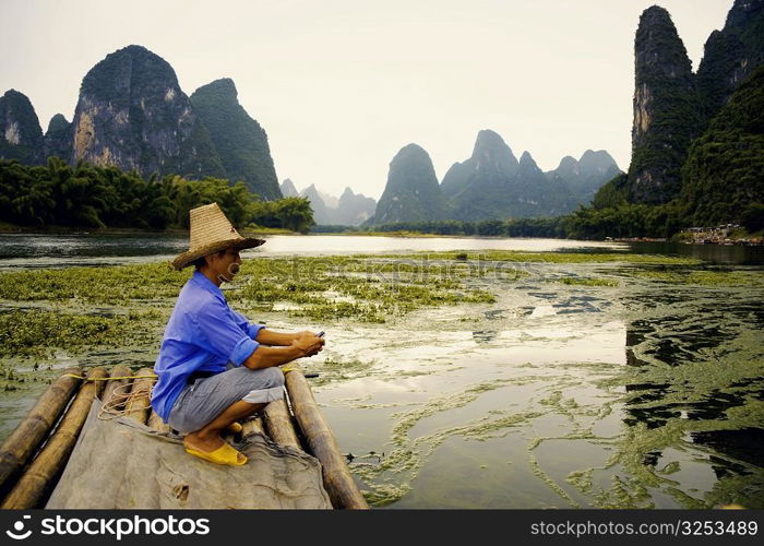 Side profile of a mature man crouching on a bamboo raft with a hill range in the background, Guilin Hills, XingPing, Yangshuo, Guangxi Province, China