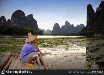 Side profile of a mature man crouching on a bamboo raft with a hill range in the background, Guilin Hills, XingPing, Yangshuo, Guangxi Province, China