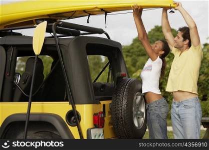 Side profile of a mid adult man and a young woman taking a kayak from the roof of a jeep