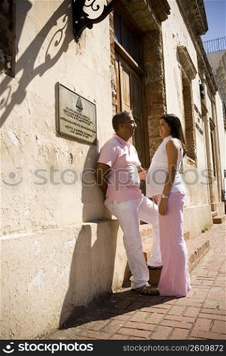 Side profile of a mid adult man leaning against a wall with a pregnant young woman standing in front of him