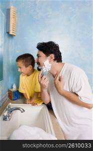 Side profile of a mid adult man shaving with his son sitting beside him in the bathroom