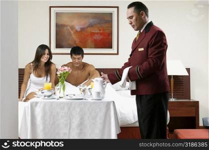 Side profile of a waiter serving a young woman and a mid adult man breakfast in bed